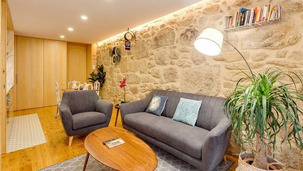 A cozy living room with a stone wall, a grey sofa, two armchairs, a wooden coffee table, and a bookshelf. - *NEW* Riverside 2 Bedroom Apartment by DA'HOME (Porto)
