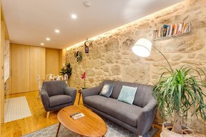A cozy living room with a stone wall, a grey sofa, two armchairs, a wooden coffee table, and a bookshelf. - *NEW* Riverside 2 Bedroom Apartment by DA'HOME (Porto)