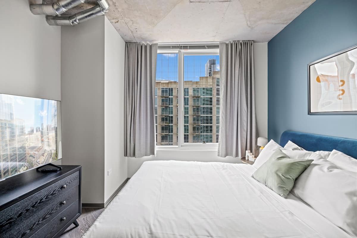 A bedroom with a large bed, a dresser, a TV on the dresser, a window with curtains, and a city view.