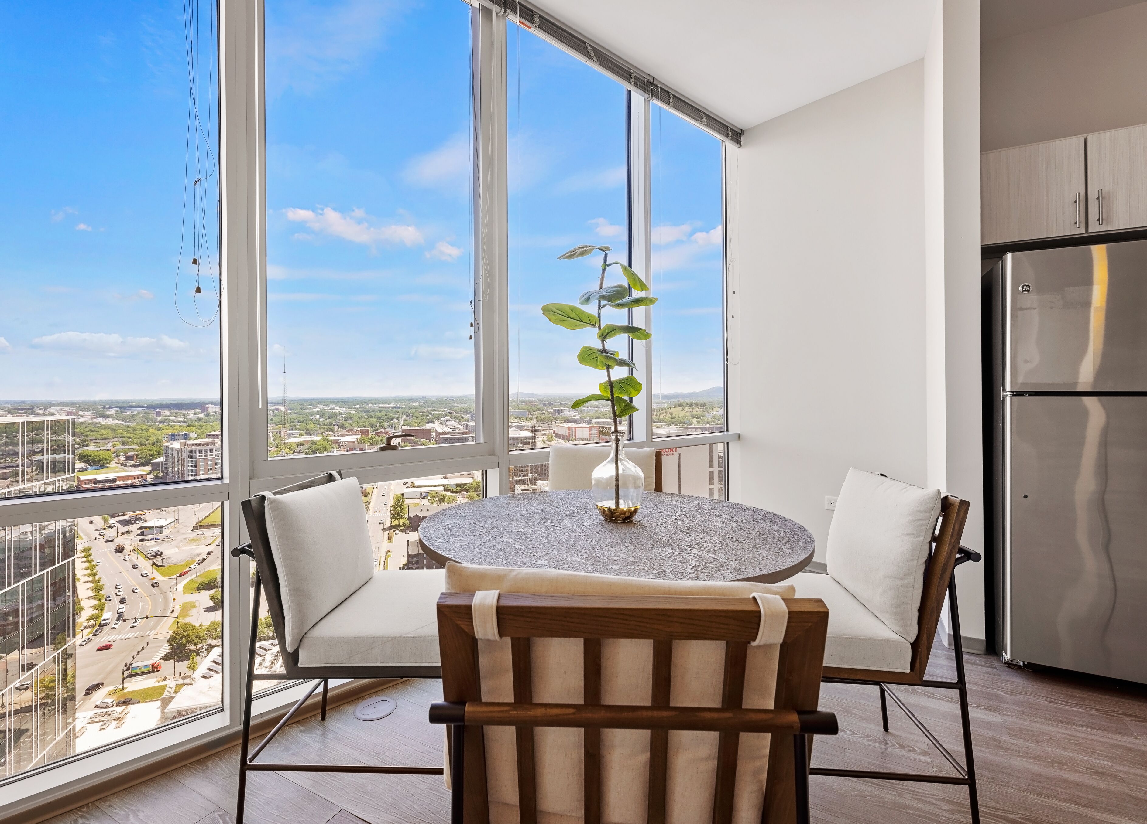 Uma varanda com mesa redonda, duas cadeiras, um vaso de planta e vista para a cidade.