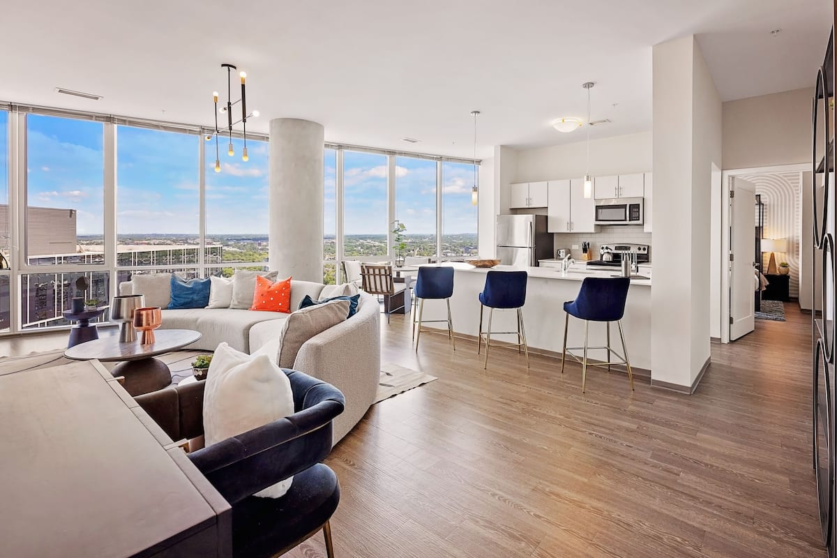 A modern apartment with a living area, kitchen, and large windows offering a city view.
