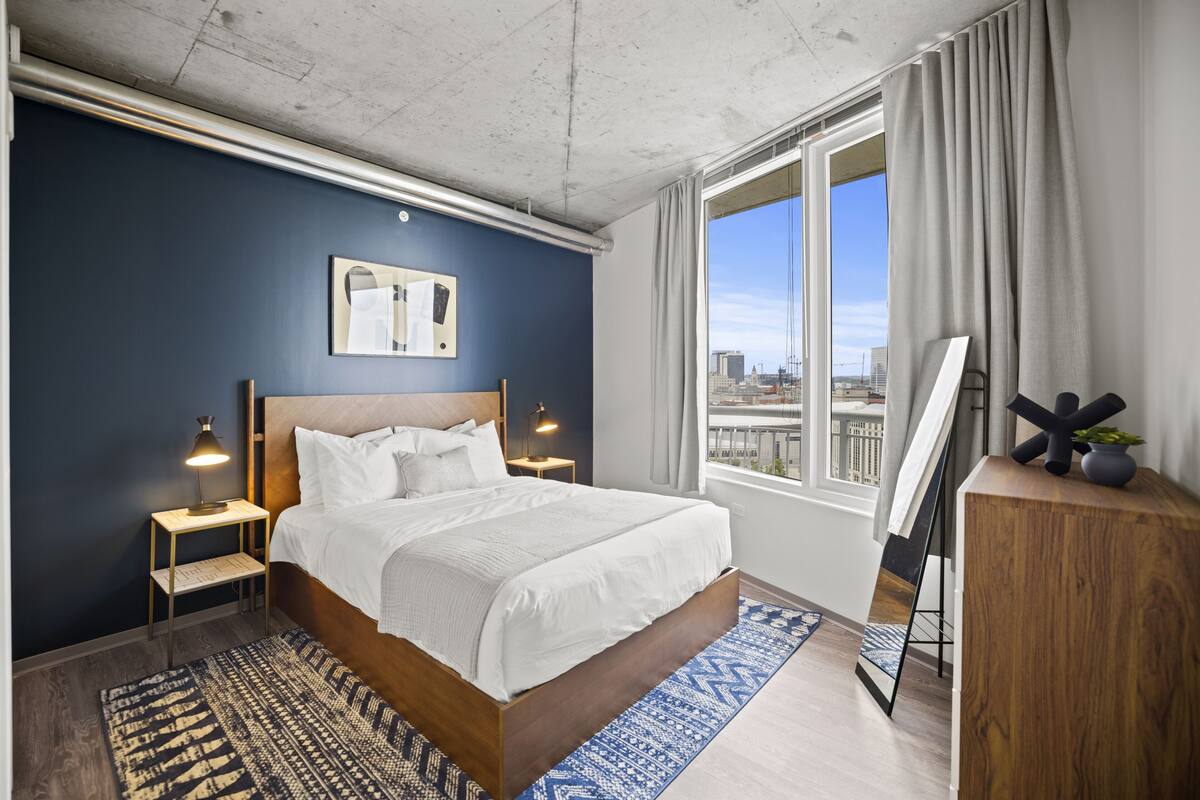 A modern hotel room with a large bed, a city view, a desk, and a standing art piece.