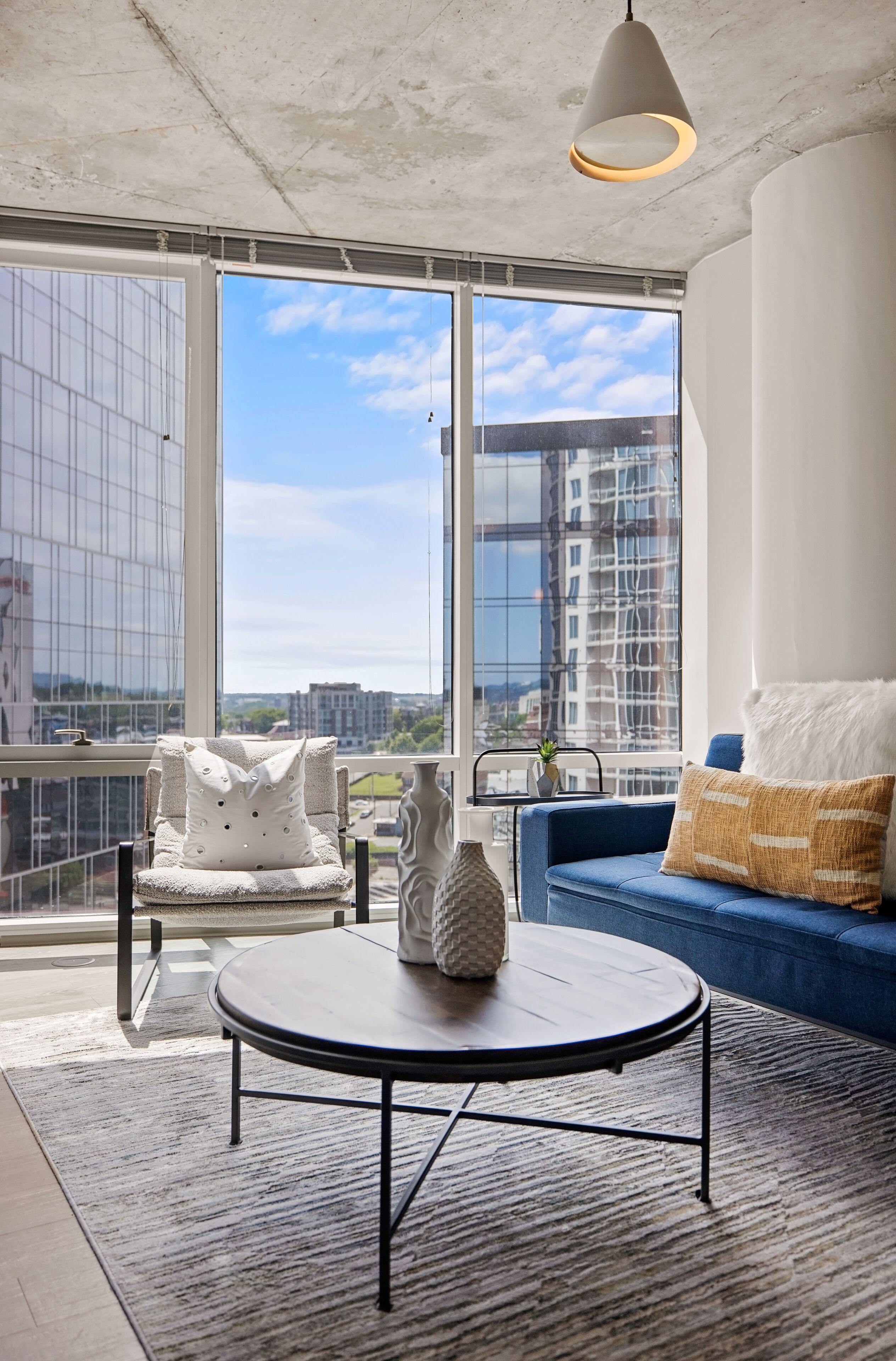 A modern living room with a round coffee table, a blue sofa, and a white armchair with a patterned cushion. Large windows offer a view of the cityscape and a building under construction.