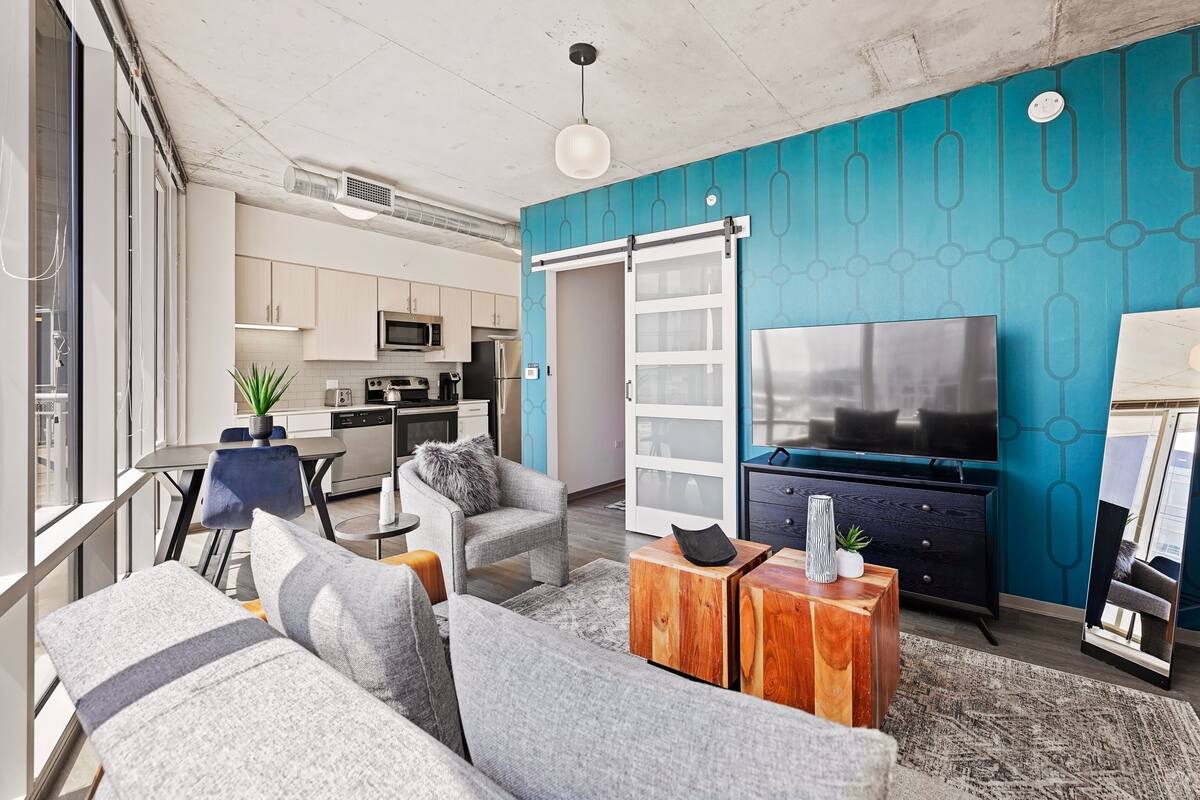 A modern living room with a sofa, coffee table, and TV. There is a kitchen area with cabinets and appliances in the background.