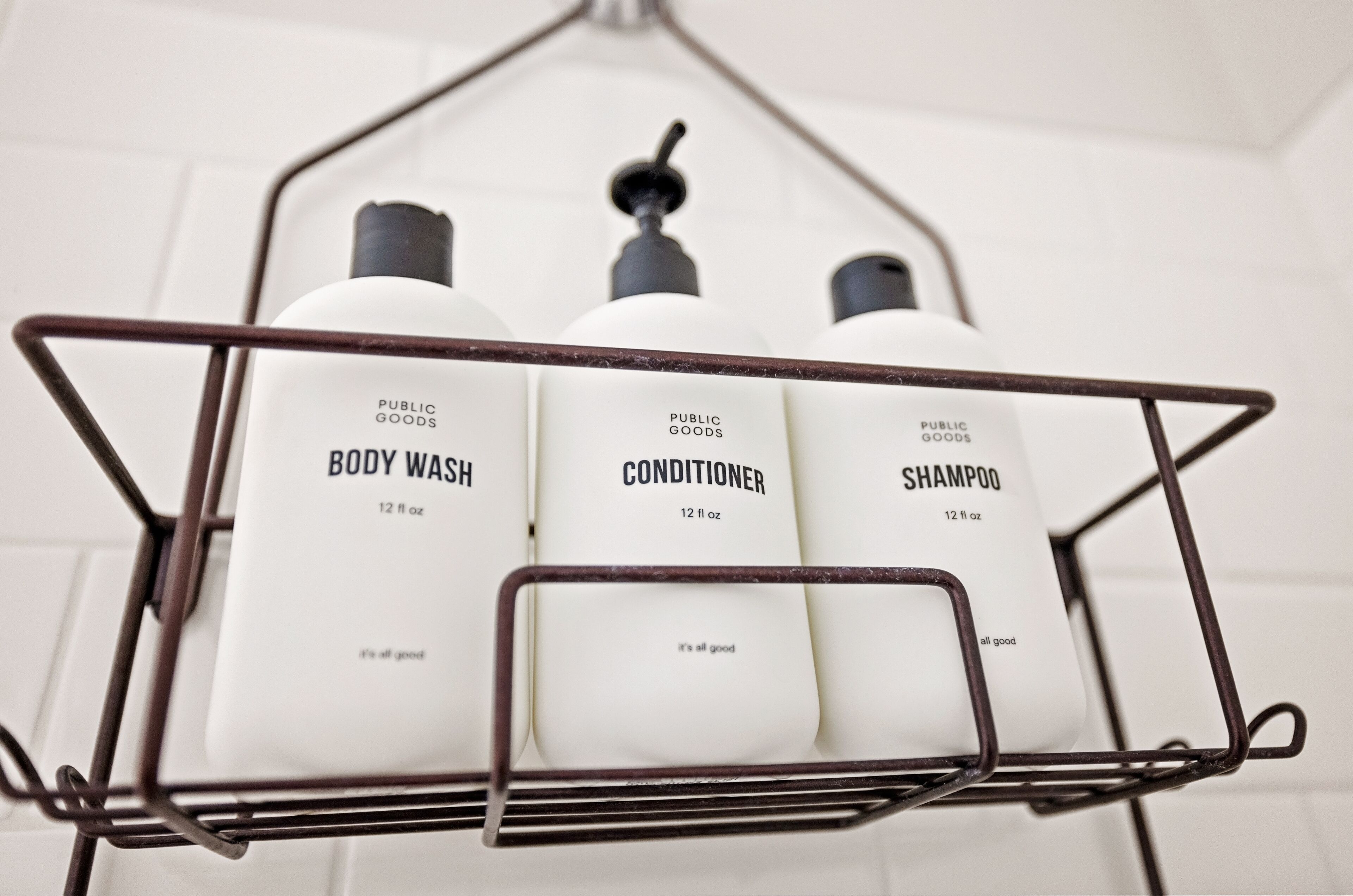 A metal rack with three white bottles labeled 'PUBLIC GOODS BODY WASH', 'PUBLIC GOODS CONDITIONER', and 'PUBLIC GOODS SHAMPOO', each 12 fl oz.