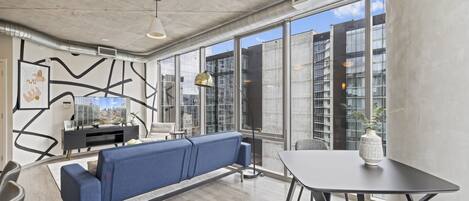 A modern interior with a blue sofa, a dining table, and large windows offering a city view.