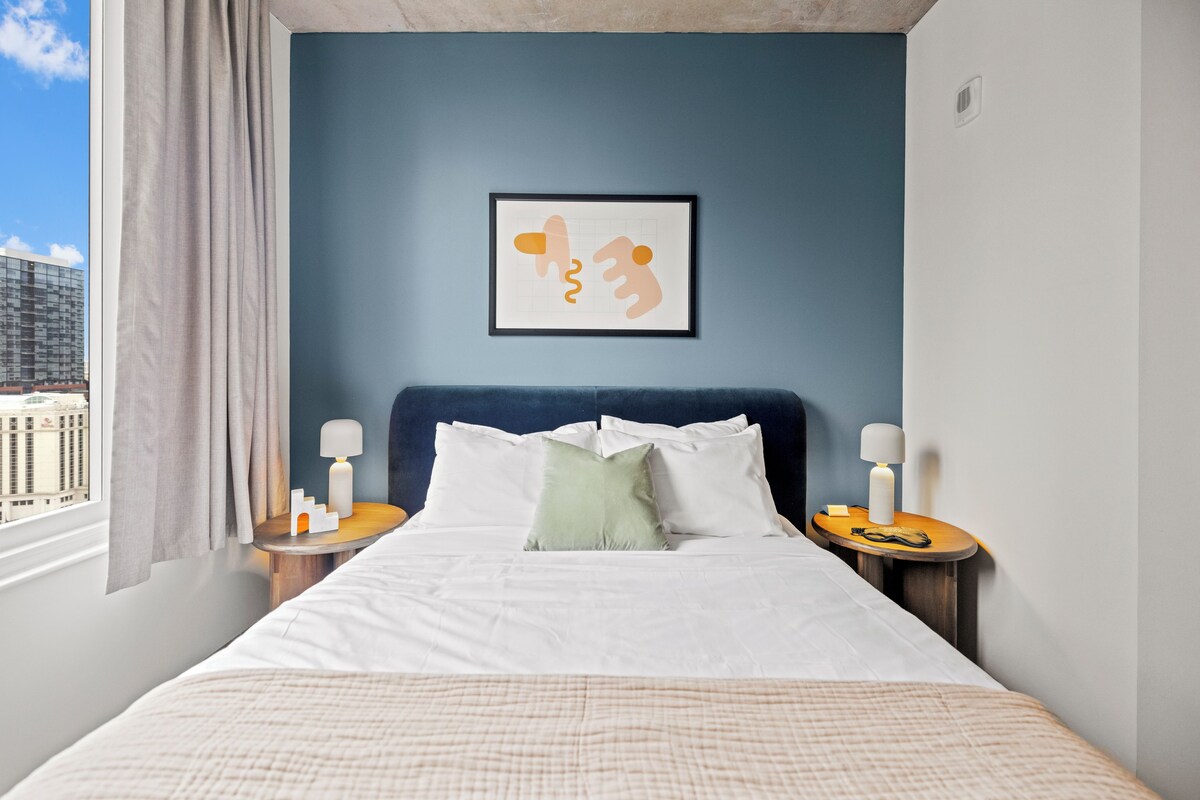 Habitación de hotel con cama, dos mesitas de noche con lámparas, un cuadro abstracto enmarcado en la pared y una ventana con cortinas.
