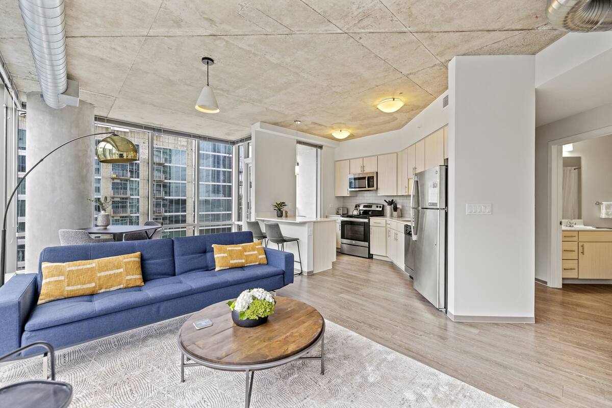 A modern open-plan living and kitchen area with a blue sofa, wooden coffee table, and large windows.