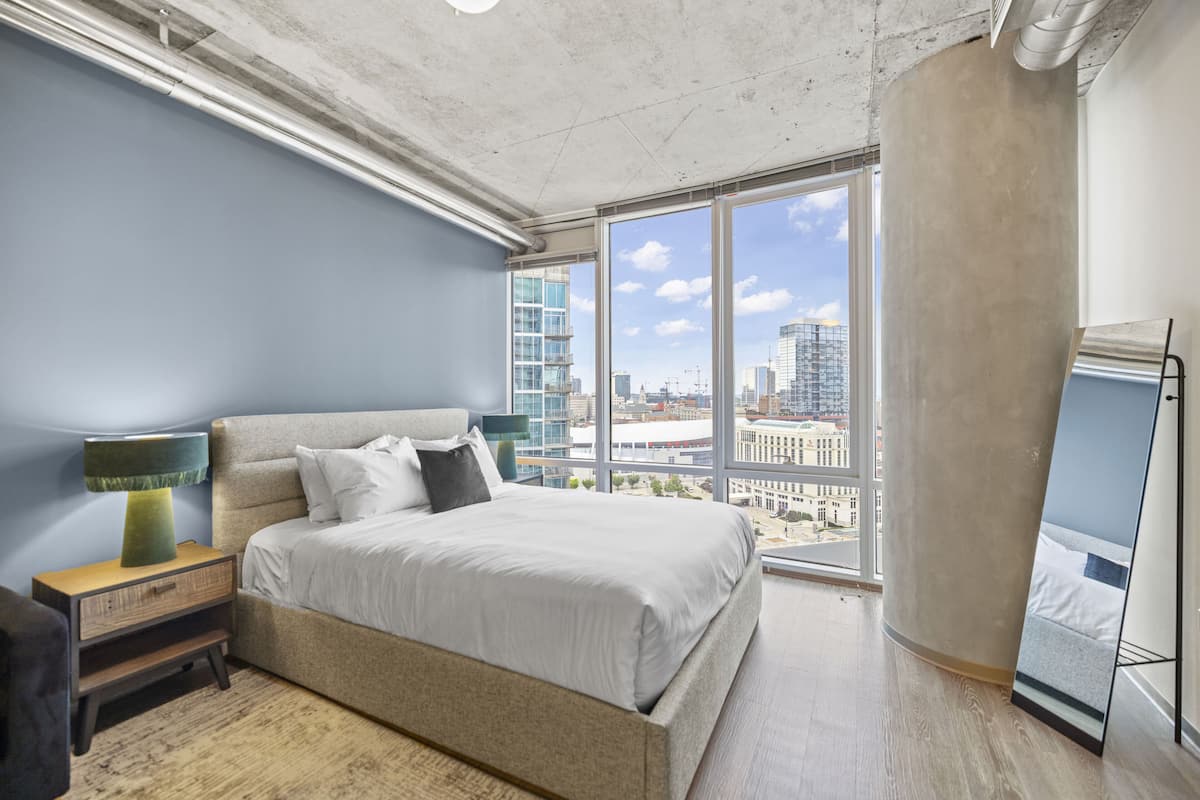 A modern bedroom with a large bed, a nightstand, a lamp, and a view of the city through large windows.