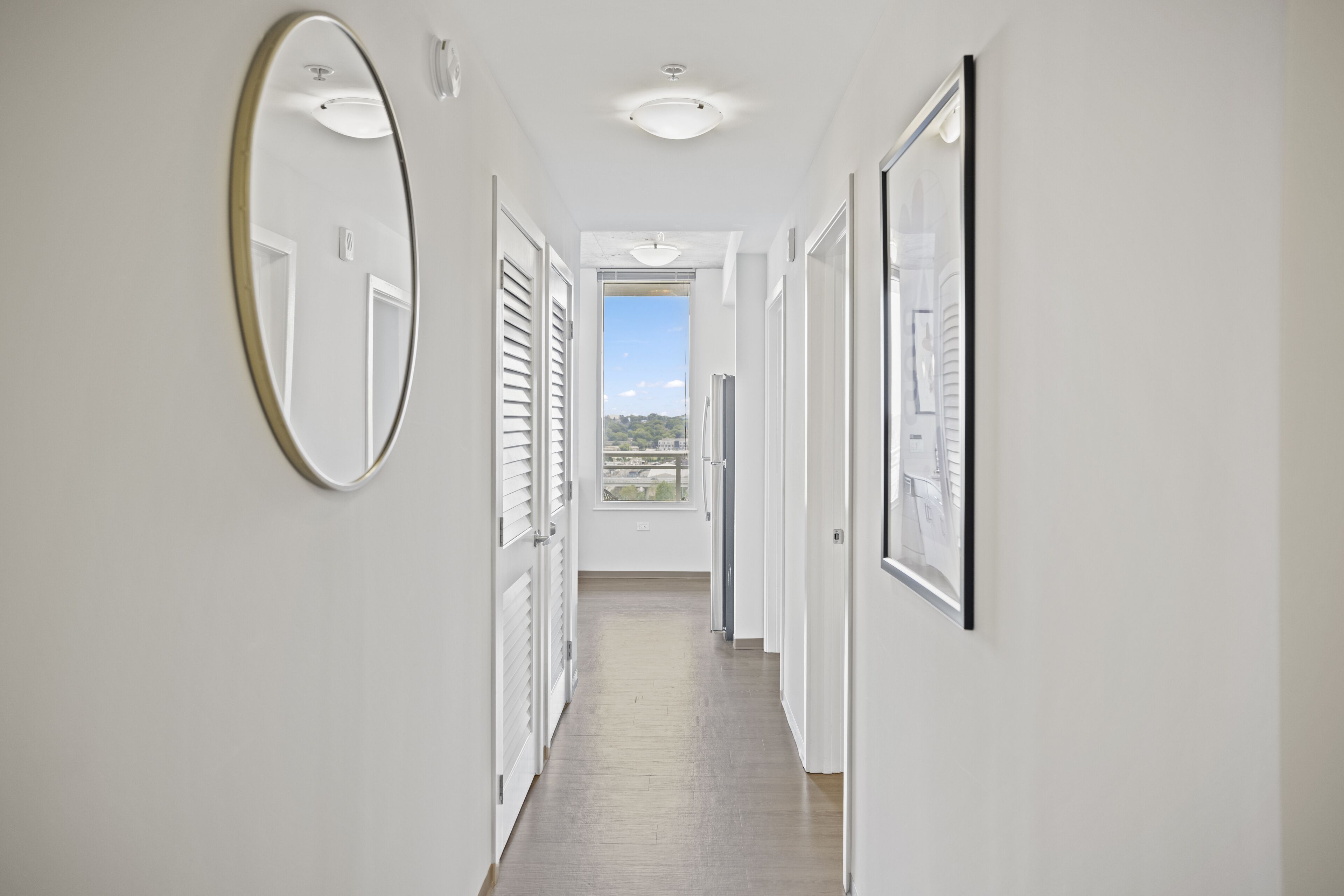 A hallway with white walls, a wooden floor, and a large window at the end.