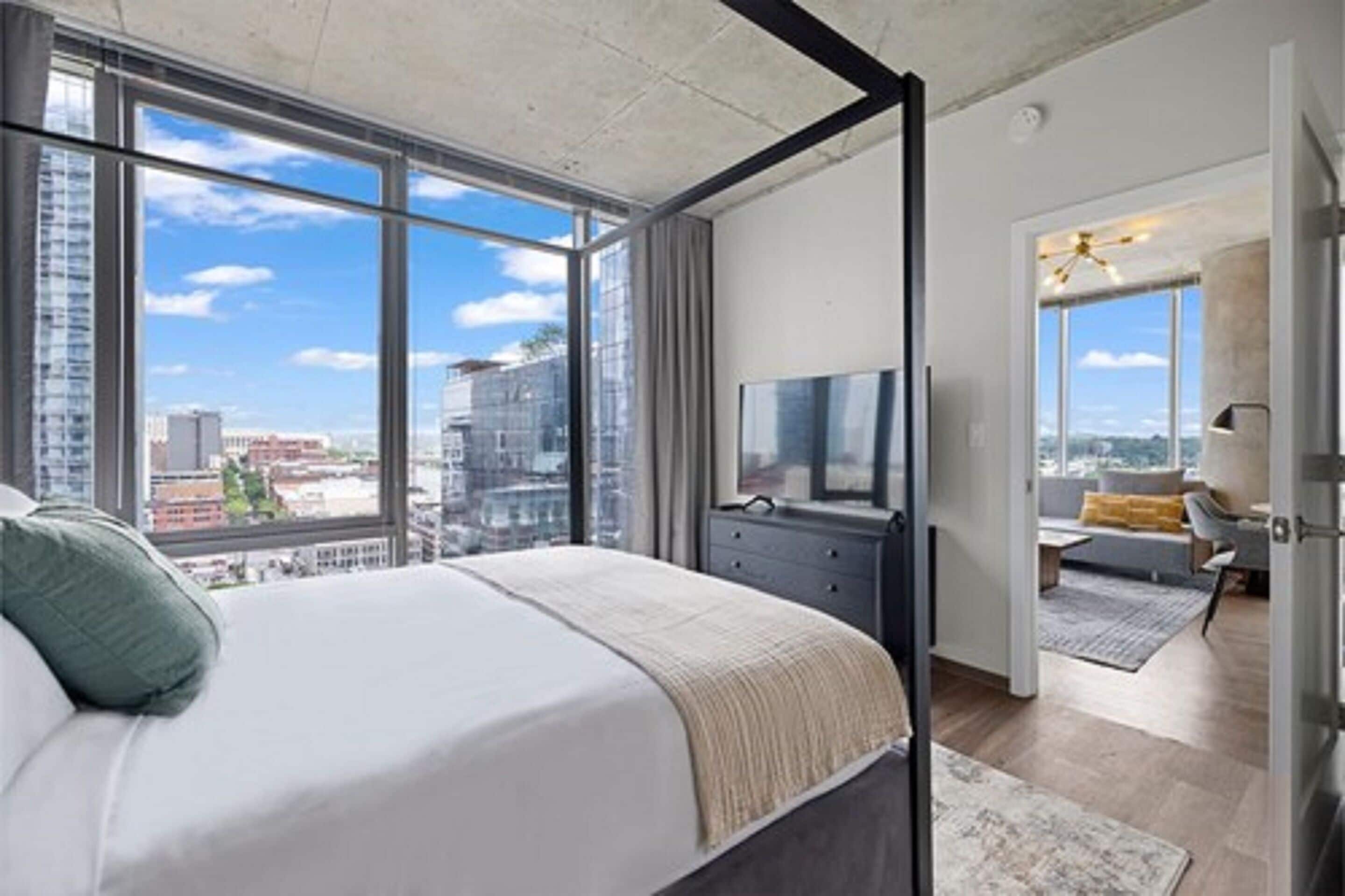 Une chambre moderne avec un grand lit, une télévision et une vue sur la ville grâce à de grandes fenêtres.