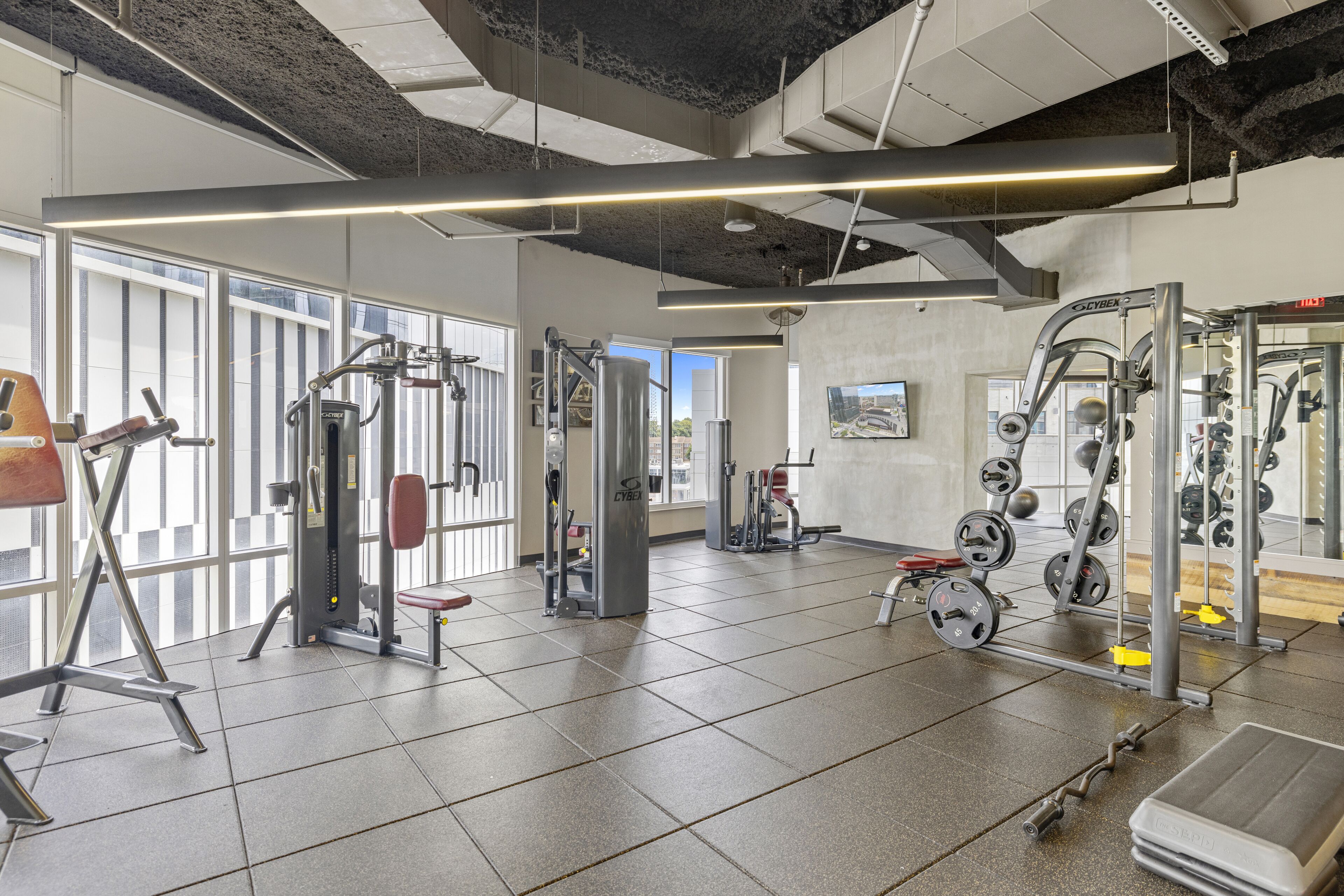 Un gimnasio con equipo de ejercicio variado, ventanas amplias y techo de hormigón.