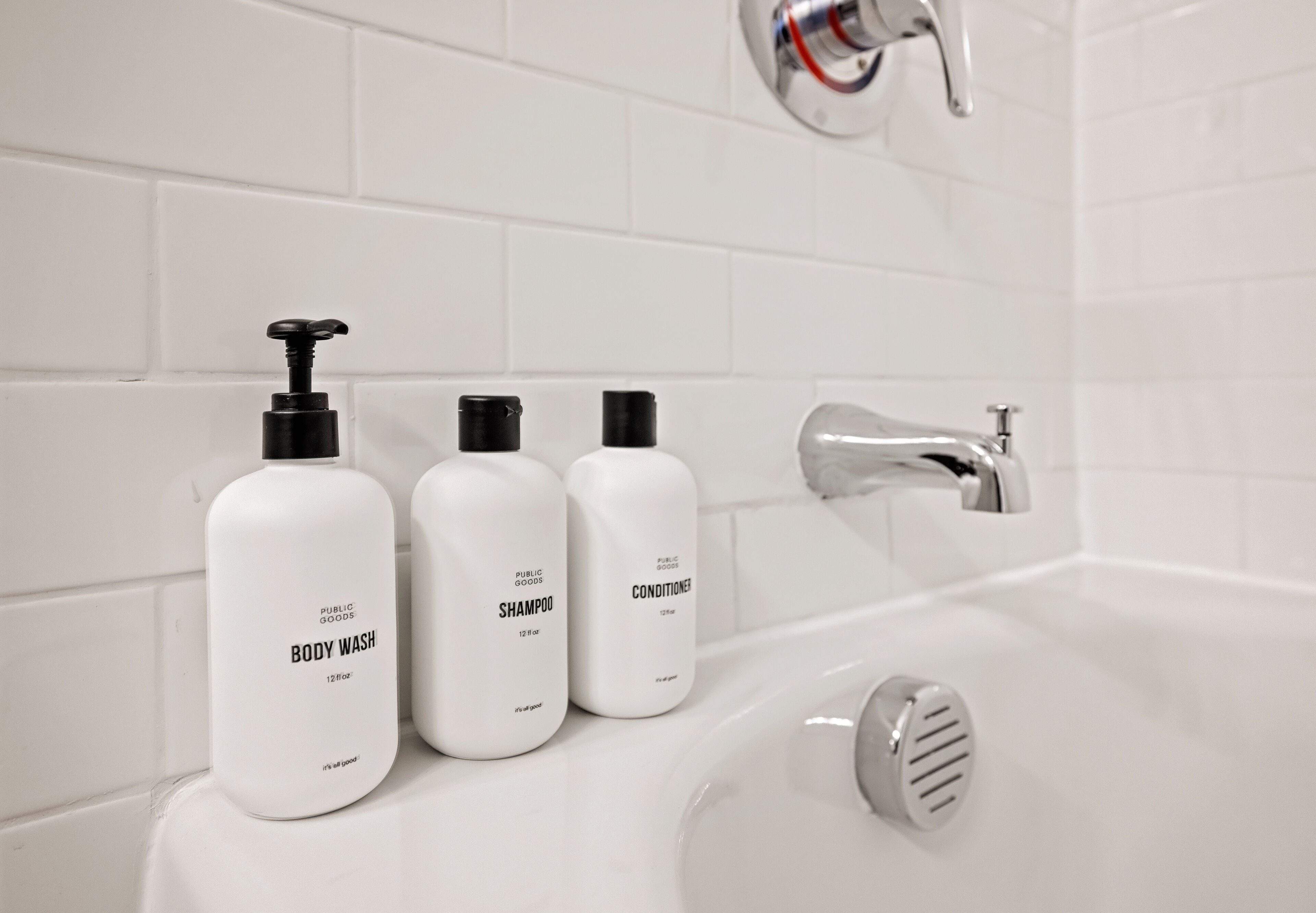 Ein Badezimmer mit weiß gefliesten Wänden, einem Chromarmaturen und drei weißen Flaschen mit den Aufschriften 'Public Goods Body Wash', 'Public Goods Shampoo' und 'Public Goods Conditioner'.