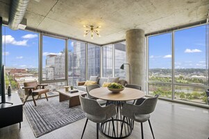Un salon moderne doté de grandes fenêtres, d’une table de salle à manger ronde et de chaises recouvertes d’un tissu gris.