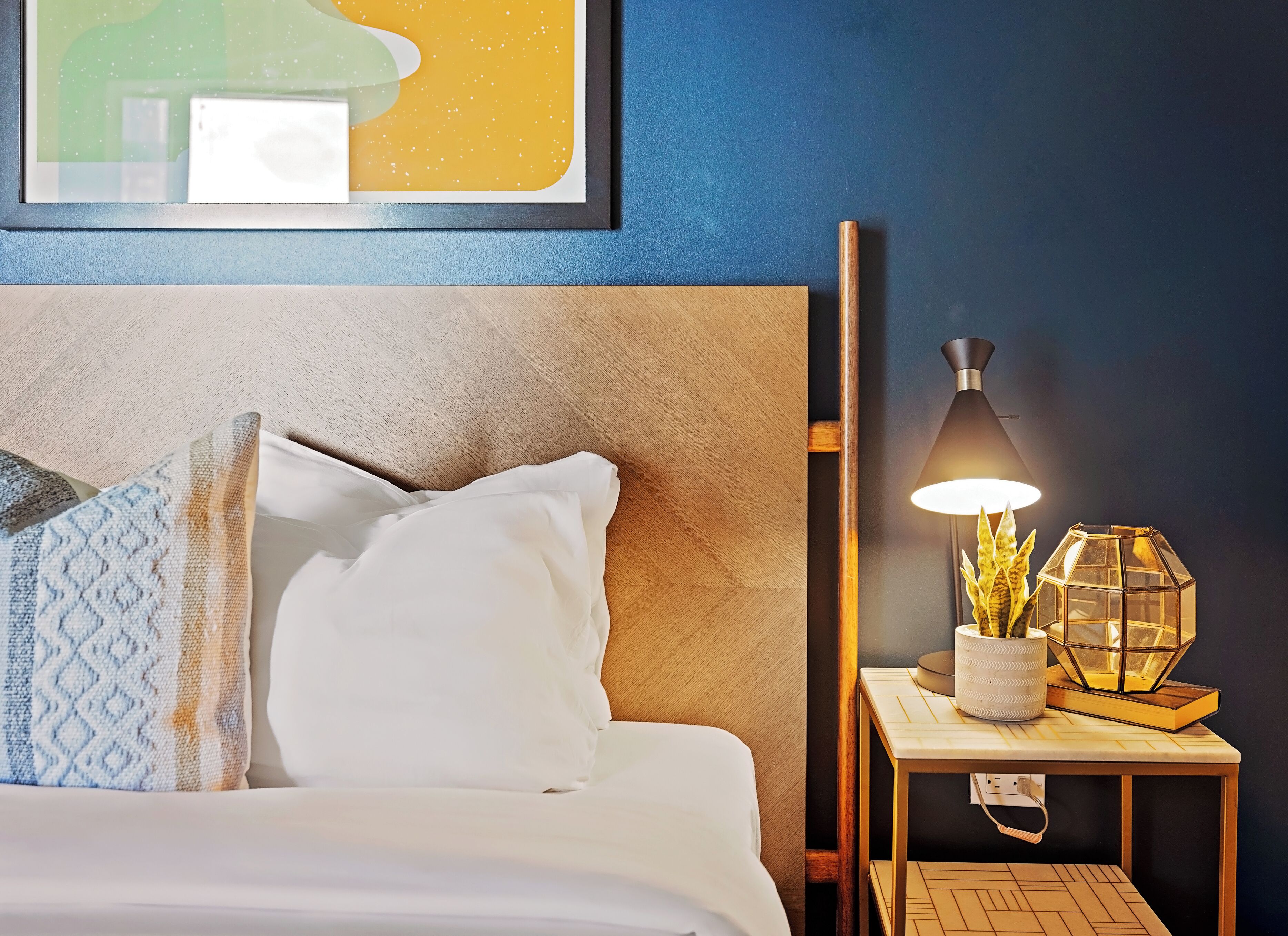 A neatly made bed with white bedding and patterned pillows, a bedside table with a lamp and decorative items, and a framed artwork on the wall.