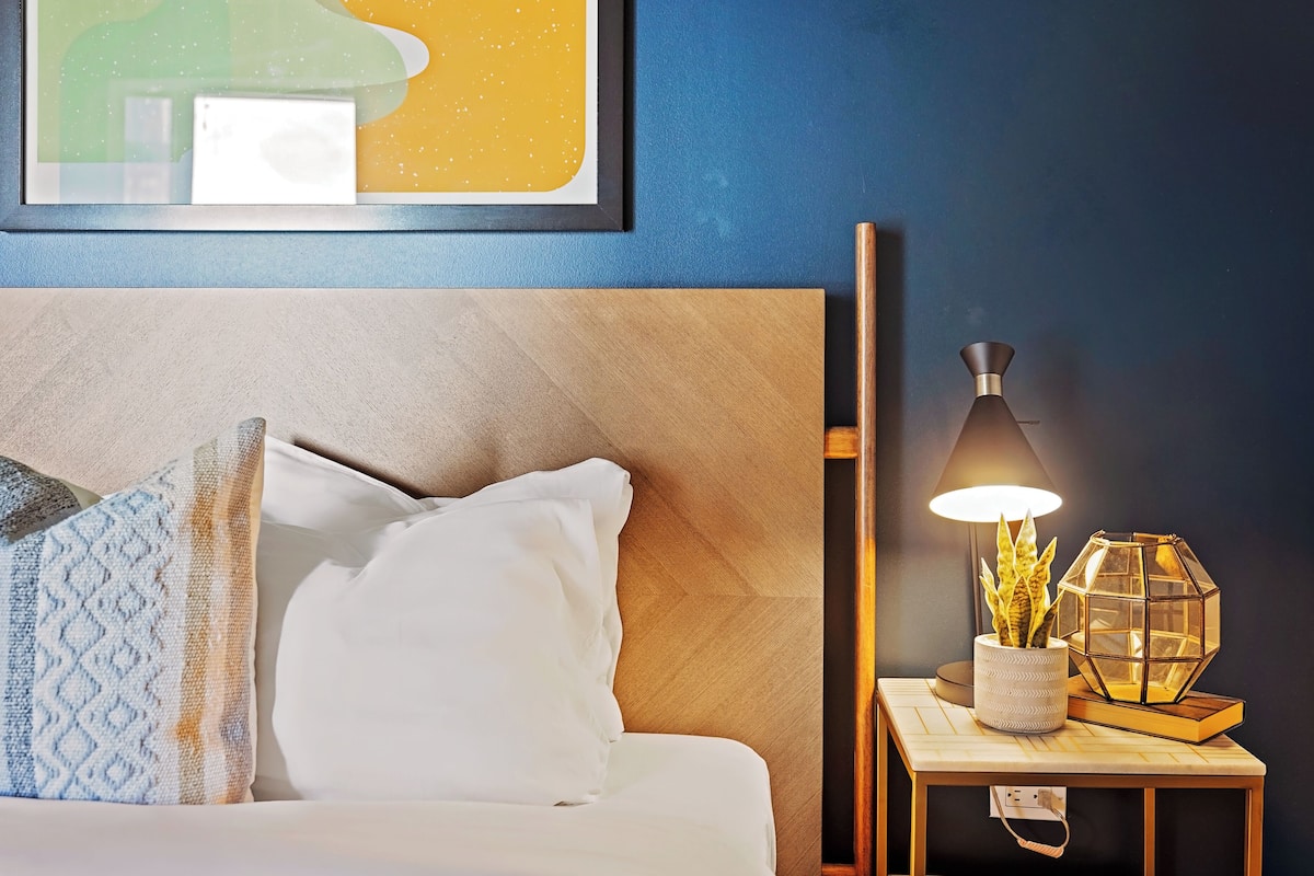 A neatly made bed with white bedding and patterned pillows, a bedside table with a lamp and decorative items, and a framed artwork on the wall.