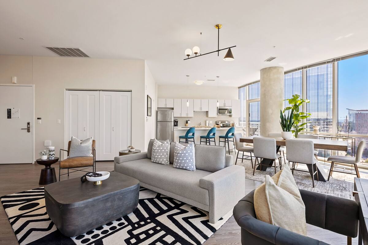 A modern living room with a grey sofa, a coffee table, and a dining area with a large window offering a city view.