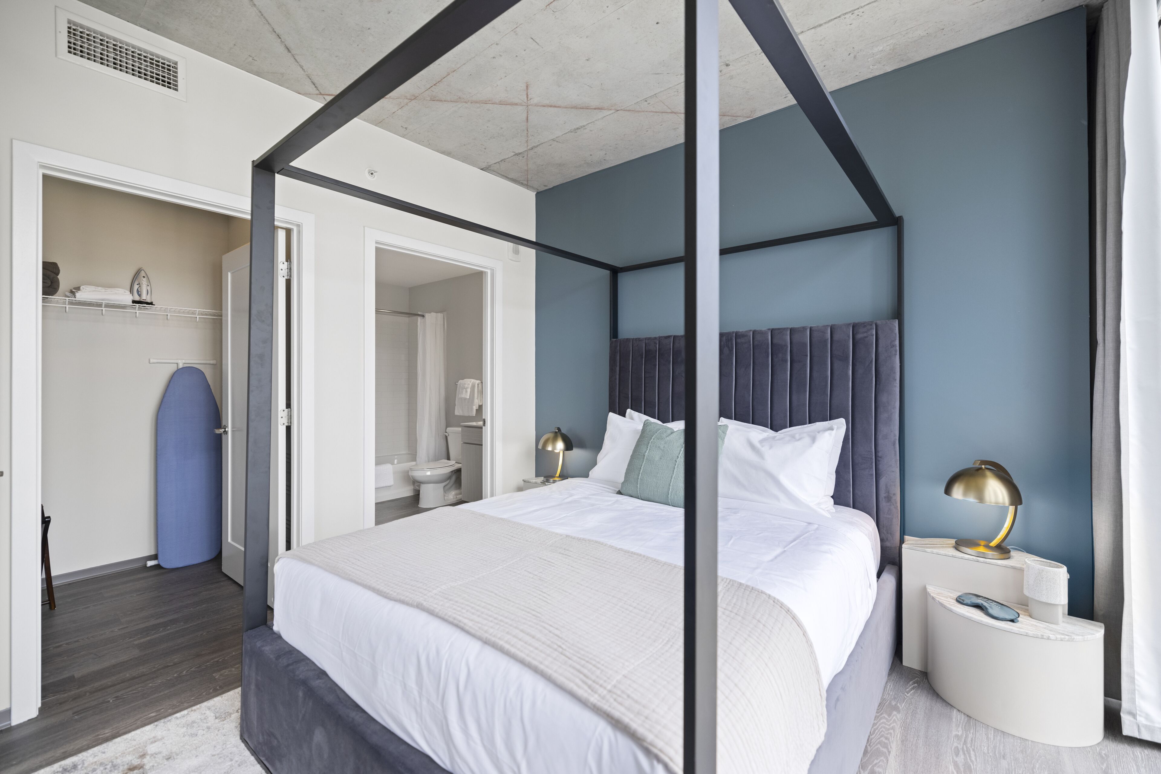 Un lit à baldaquin avec du linge de lit blanc, une table de chevet avec une lampe, et une vue sur la salle de bain et le placard.