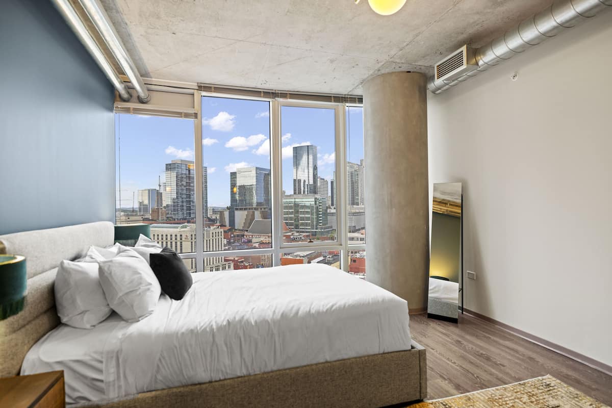 A modern hotel room with a large bed, a view of the cityscape, and a concrete pillar.