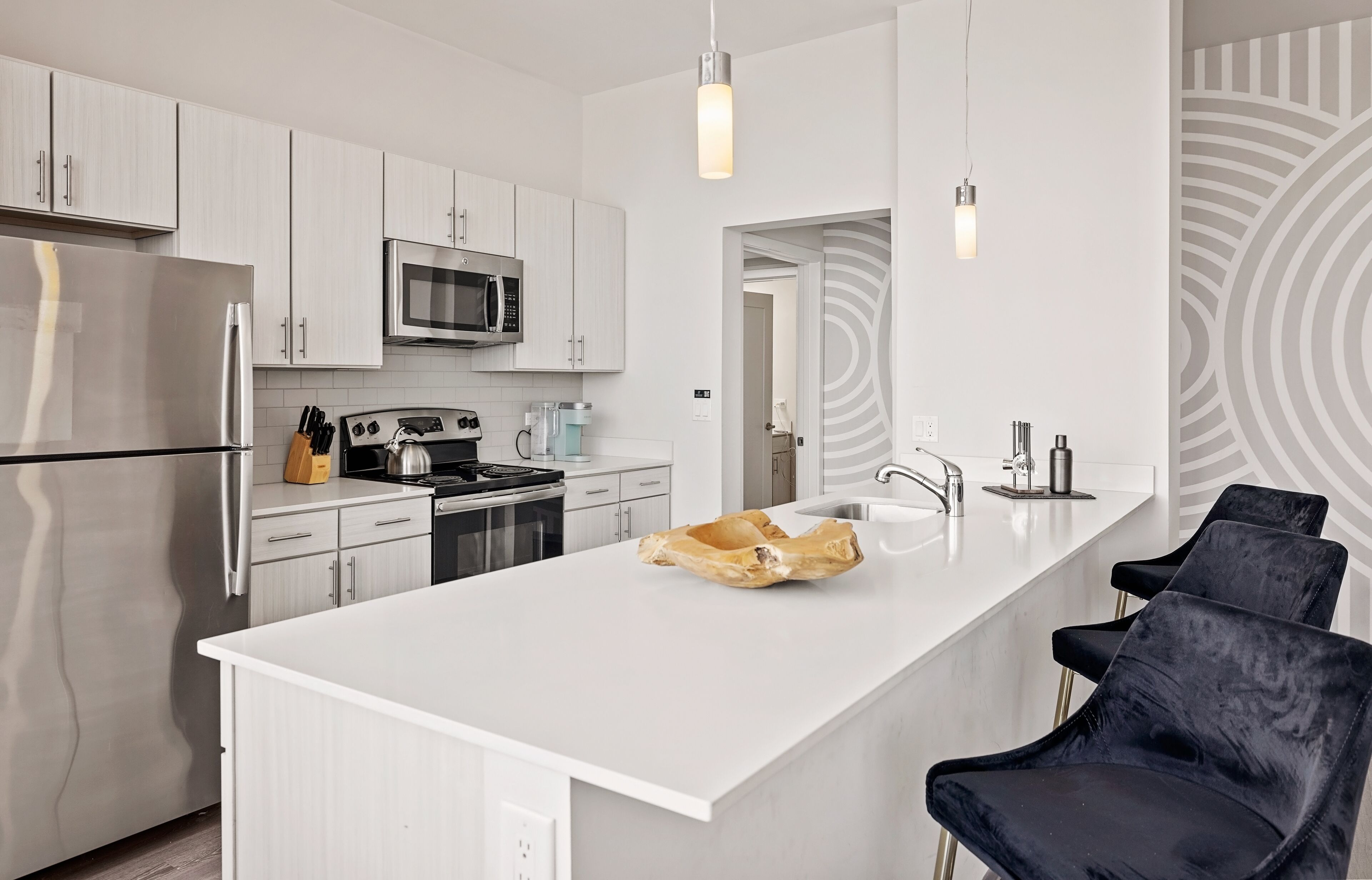 Una cocina moderna con gabinetes blancos, electrodomésticos de acero inoxidable, una isla central con taburetes altos y una pared de acento con diseño.