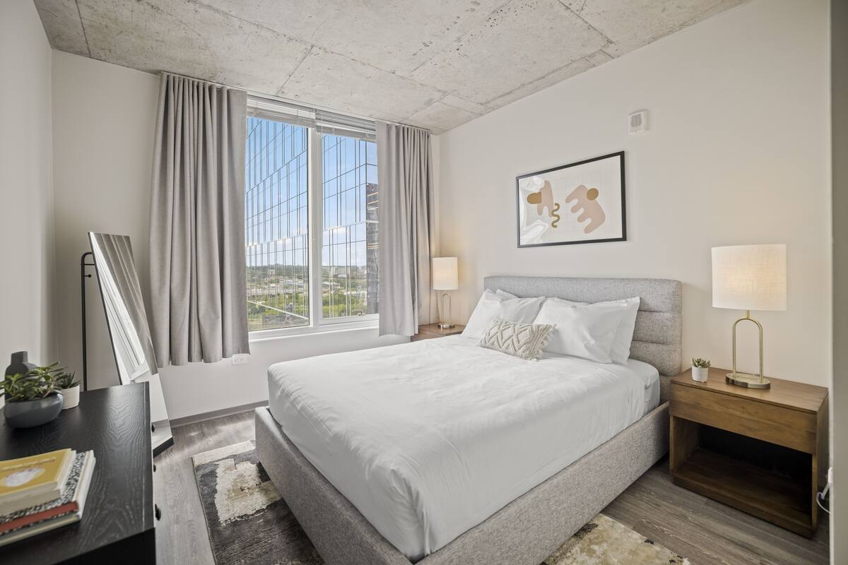 A modern hotel room with a large bed, a desk with a lamp, a framed picture on the wall, and a window with curtains.