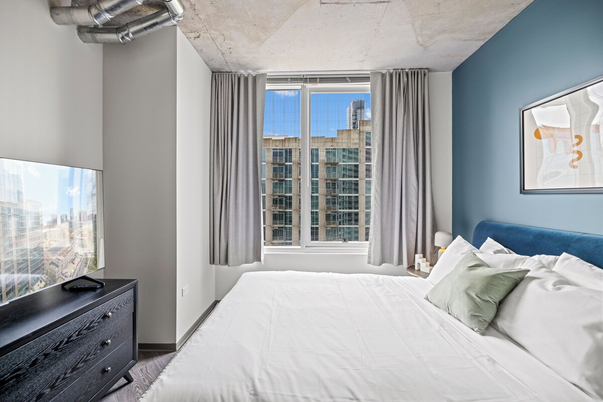 A bedroom with a large bed, a dresser, a TV on the dresser, a window with curtains, and a city view.