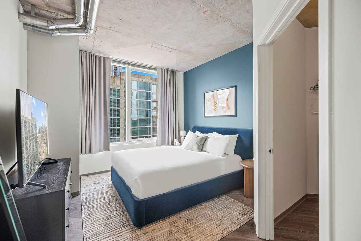 Un dormitorio con una cama grande, un televisor sobre un mueble, una ventana con cortinas y una pared azul decorativa.