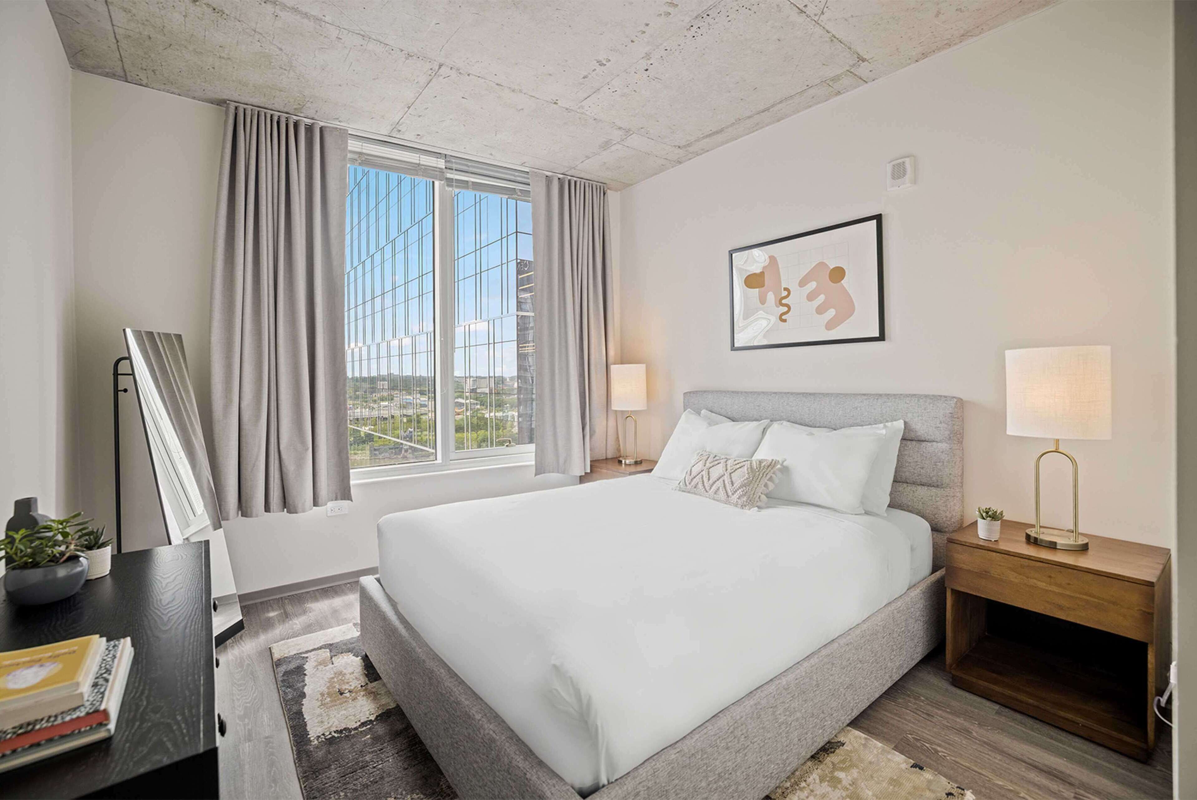 A hotel room with a large bed, a bedside table with a lamp, a framed picture on the wall, and a window with curtains.