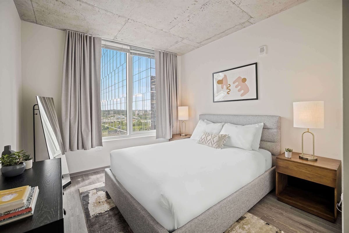 A hotel room with a large bed, a bedside table with a lamp, a framed picture on the wall, and a window with curtains.