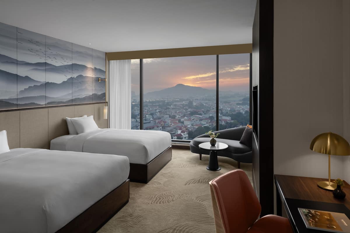 A hotel room with two beds, a sofa, a small table, and a view of a cityscape and mountains.