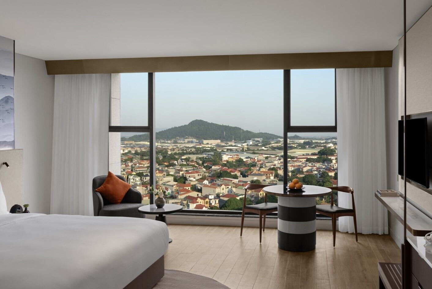 Una habitación de hotel moderna con una cama grande, una zona de estar con una mesa y vistas a la ciudad a través de grandes ventanales.