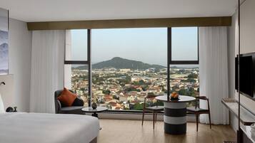 Una habitación de hotel moderna con una cama grande, una zona de estar con una mesa y vistas a la ciudad a través de grandes ventanales.