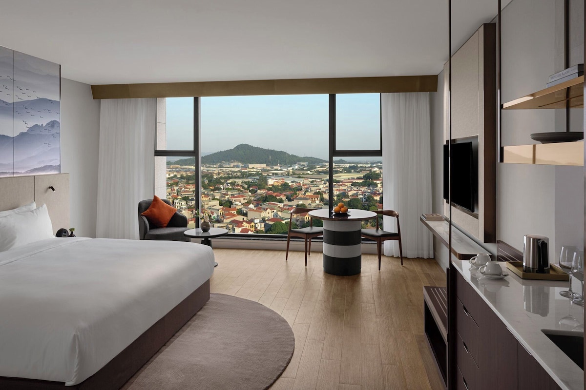 A hotel room with a large bed, a seating area with a table, a TV, and a view of a cityscape.