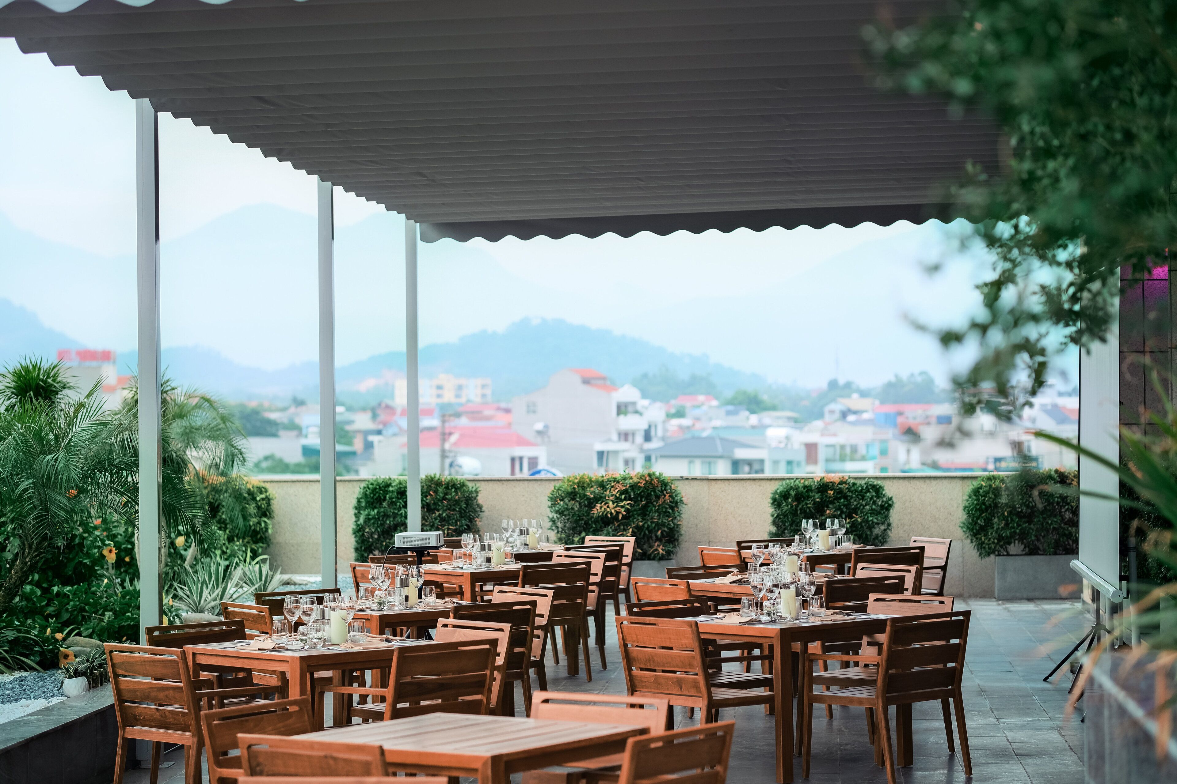 A restaurant with outdoor seating, tables set with glasses and cutlery, and a view of buildings and mountains in the background.