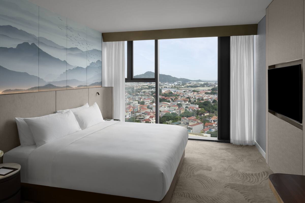 A hotel room with a large bed, a view of a cityscape through a large window, and a flat-screen TV mounted on the wall.
