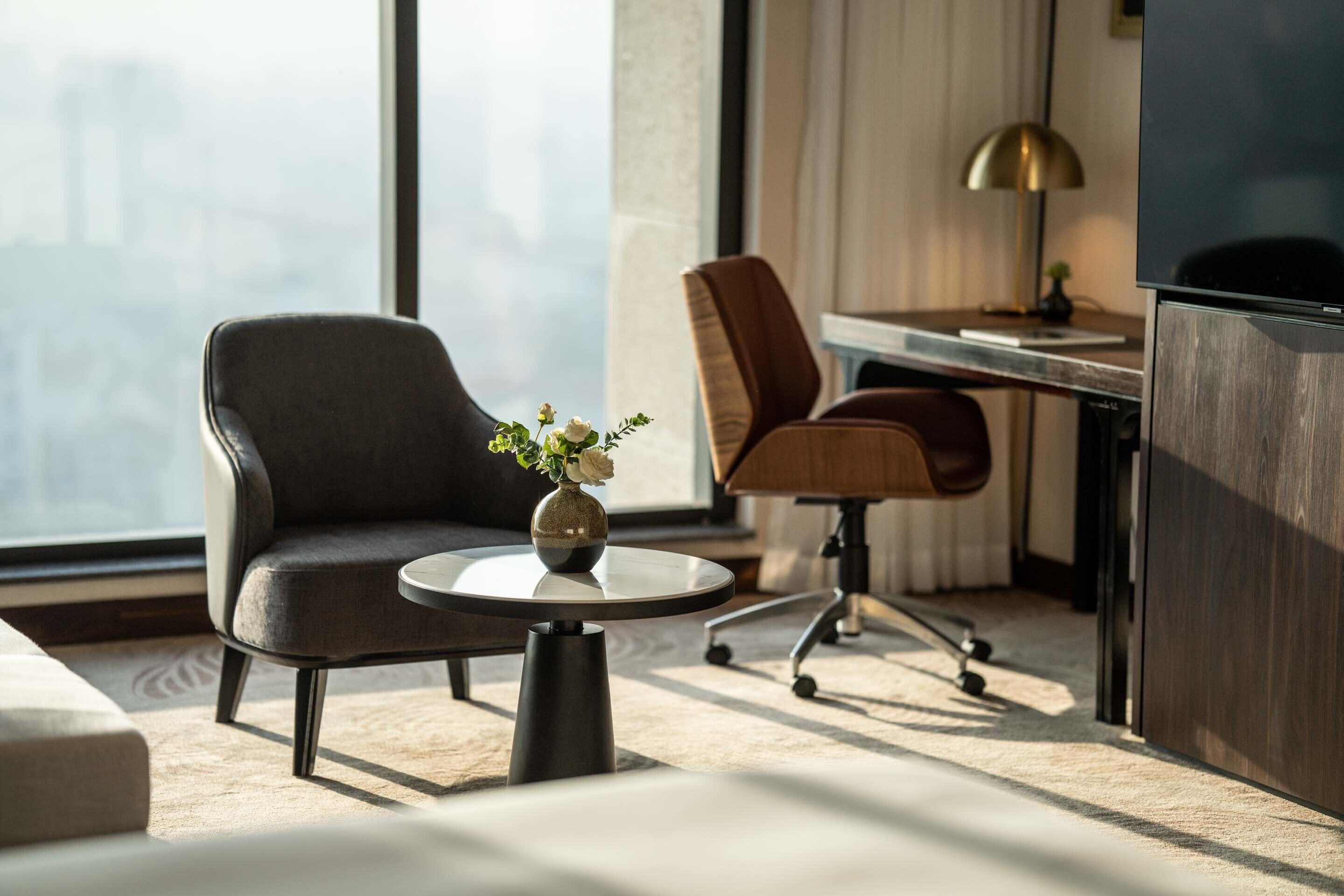 A modern hotel room with a large window, a sofa, a small round table with a vase, and a desk with a lamp.
