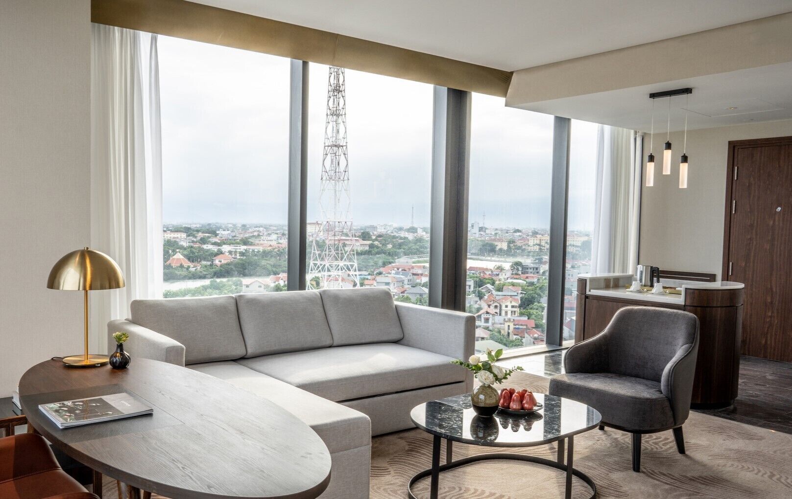 A modern hotel room with a large window, a sofa, a chair, a small table, and a desk.