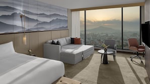 Ett modernt hotellrum med en stor säng, en grå soffa, ett litet bord med en vas och utsikt över en stadssiluett genom stora fönster.