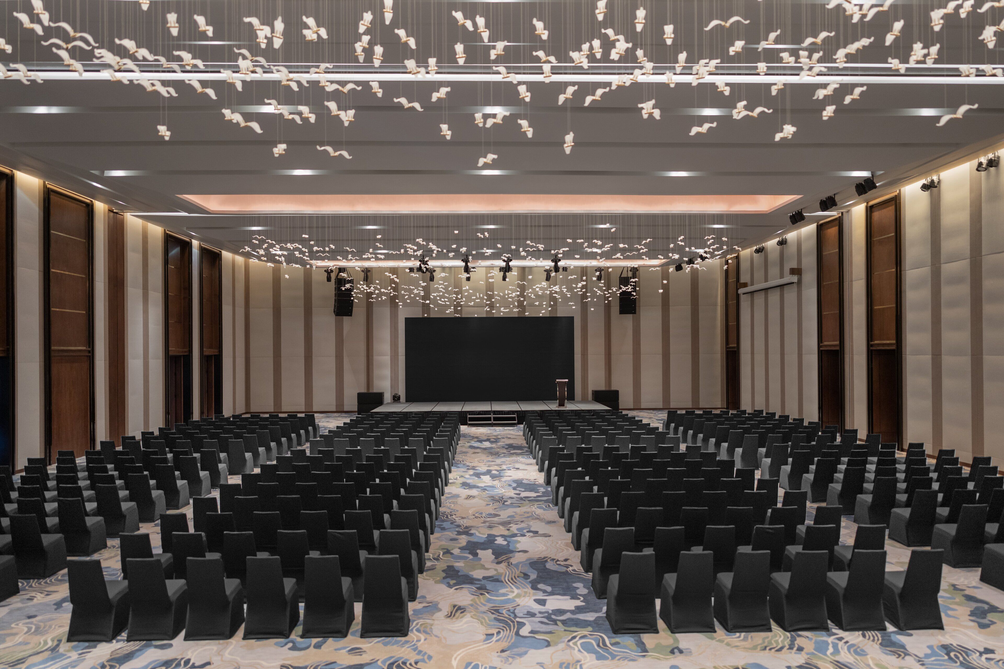 Une salle de conférence avec des rangées de chaises noires, une scène dotée d’un grand écran et un plafond orné de luminaires suspendus.