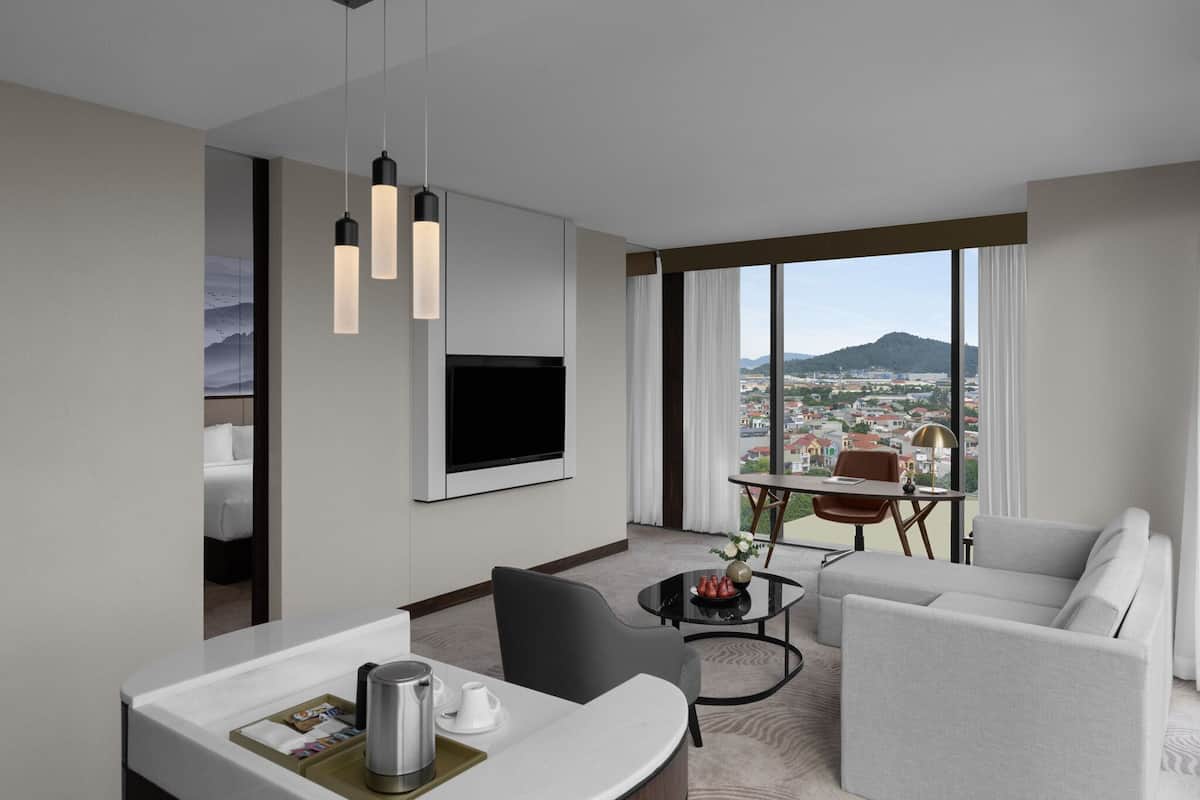 A modern hotel room with a large window offering a city view, a flat-screen TV mounted on the wall, a dining area with a table and chairs, and a sofa.