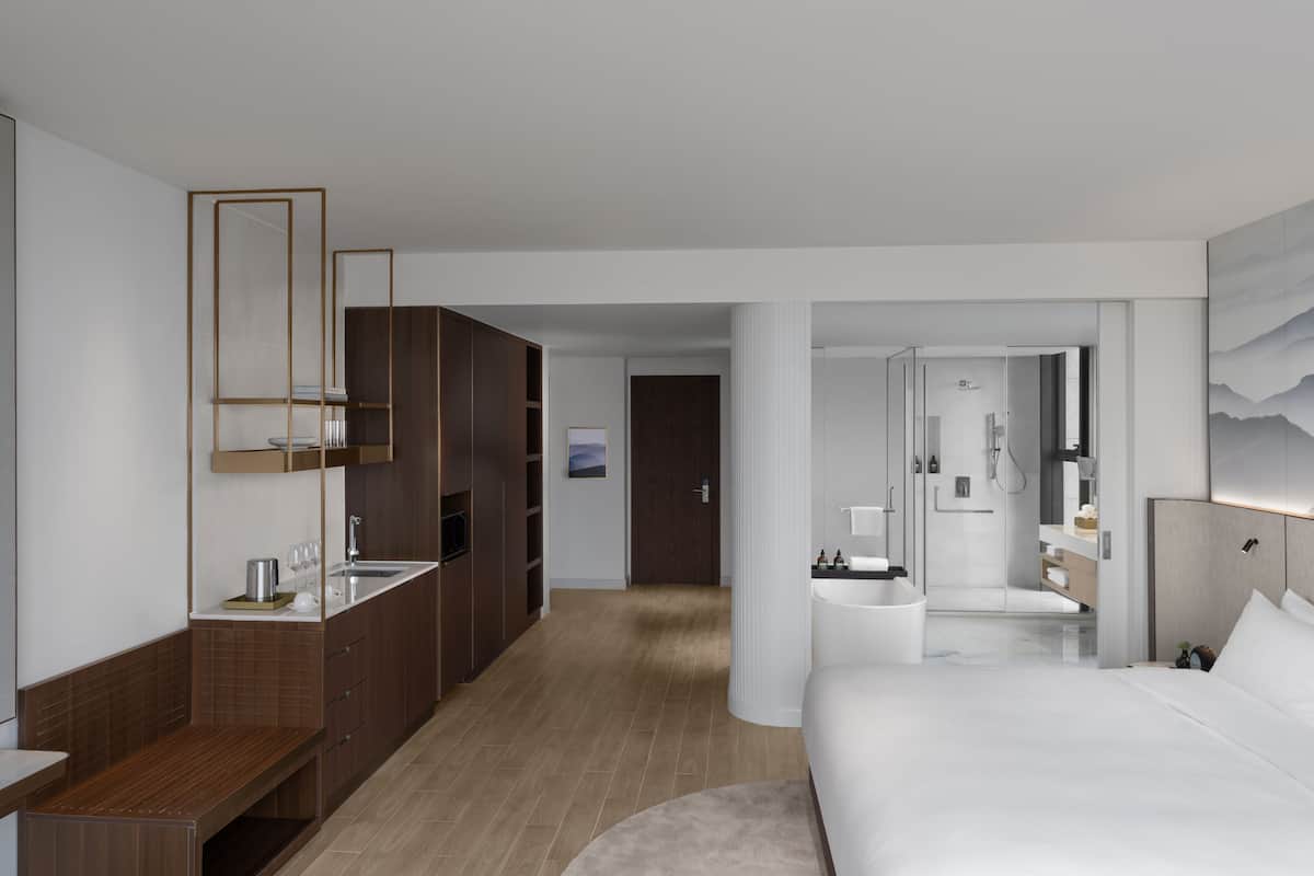 A modern hotel room with a wooden vanity, a large bed, a separate bathroom with a shower, and a view of the hallway.