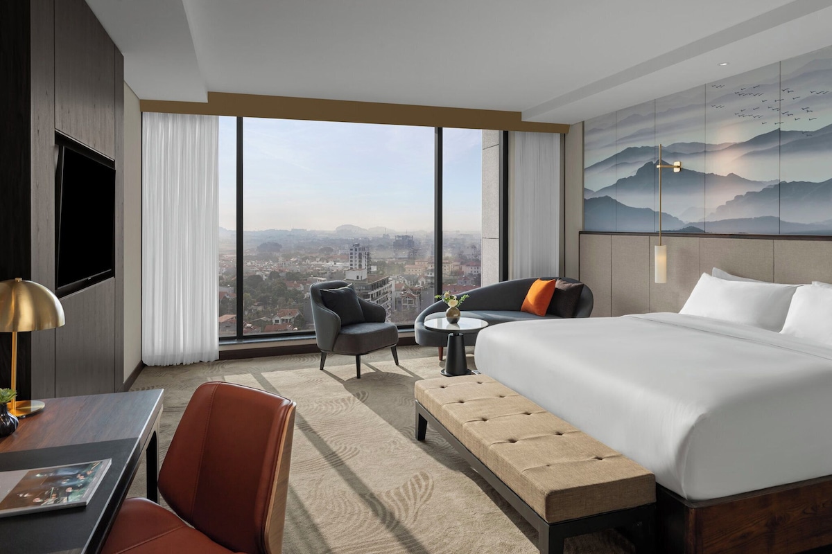 A hotel room with a large window, a bed, a desk, and a chair.