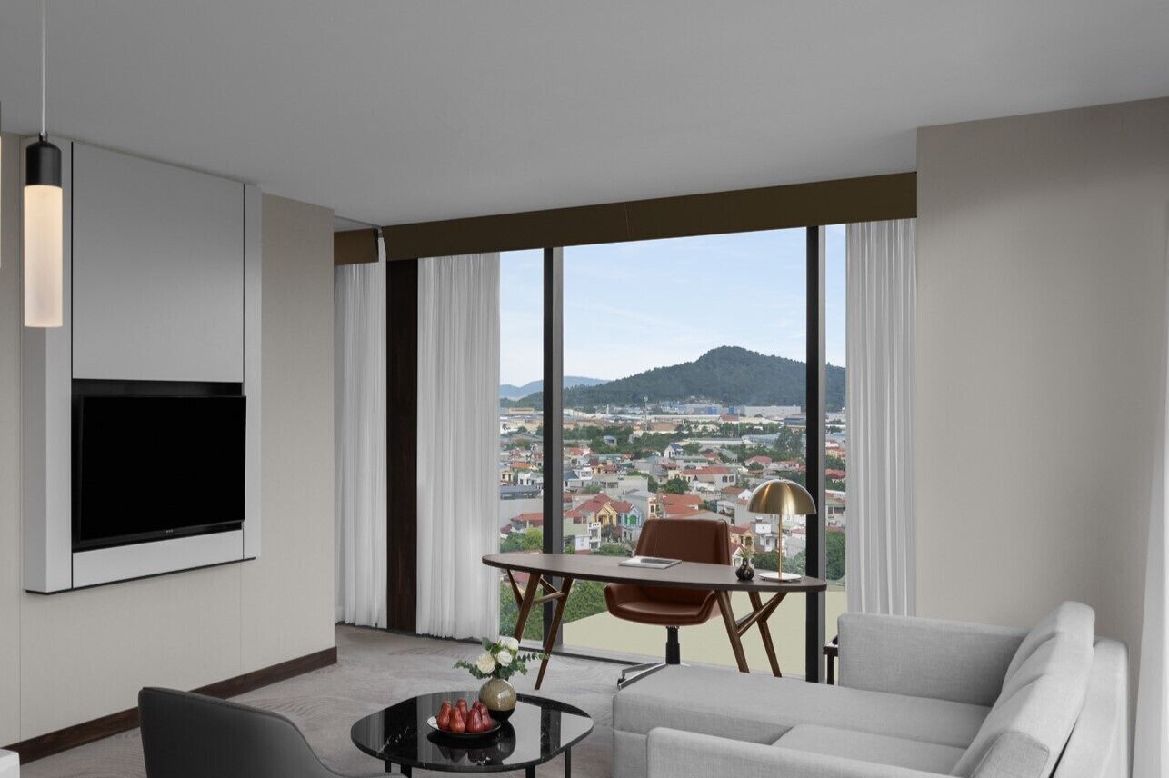 A modern hotel room with a large window offering a city view, a flat-screen TV mounted on the wall, a sofa, a dining table with chairs, and a small side table with a lamp.