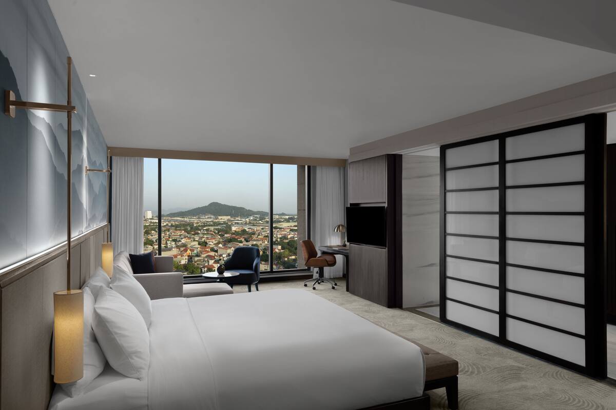A modern hotel room with a large bed, a desk, a chair, and a view of a cityscape through floor-to-ceiling windows.