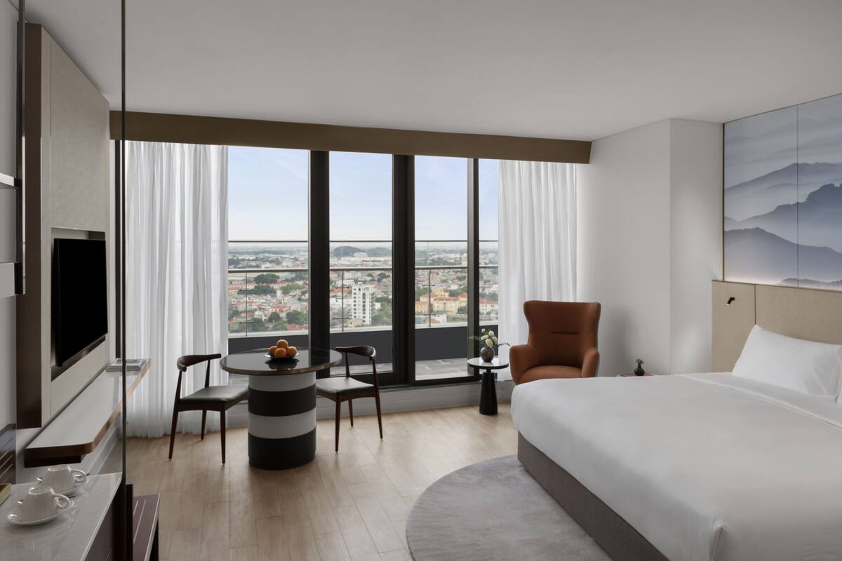 A modern hotel room with a large bed, a TV, a dining area with a table and chairs, and a view of the city through large windows.