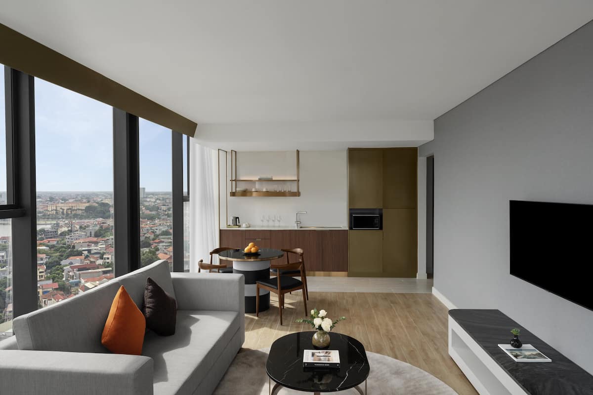 A modern living room with a sofa, coffee table, and a dining area with a table and chairs. A TV is mounted on the wall, and there is a view of the city through large windows.