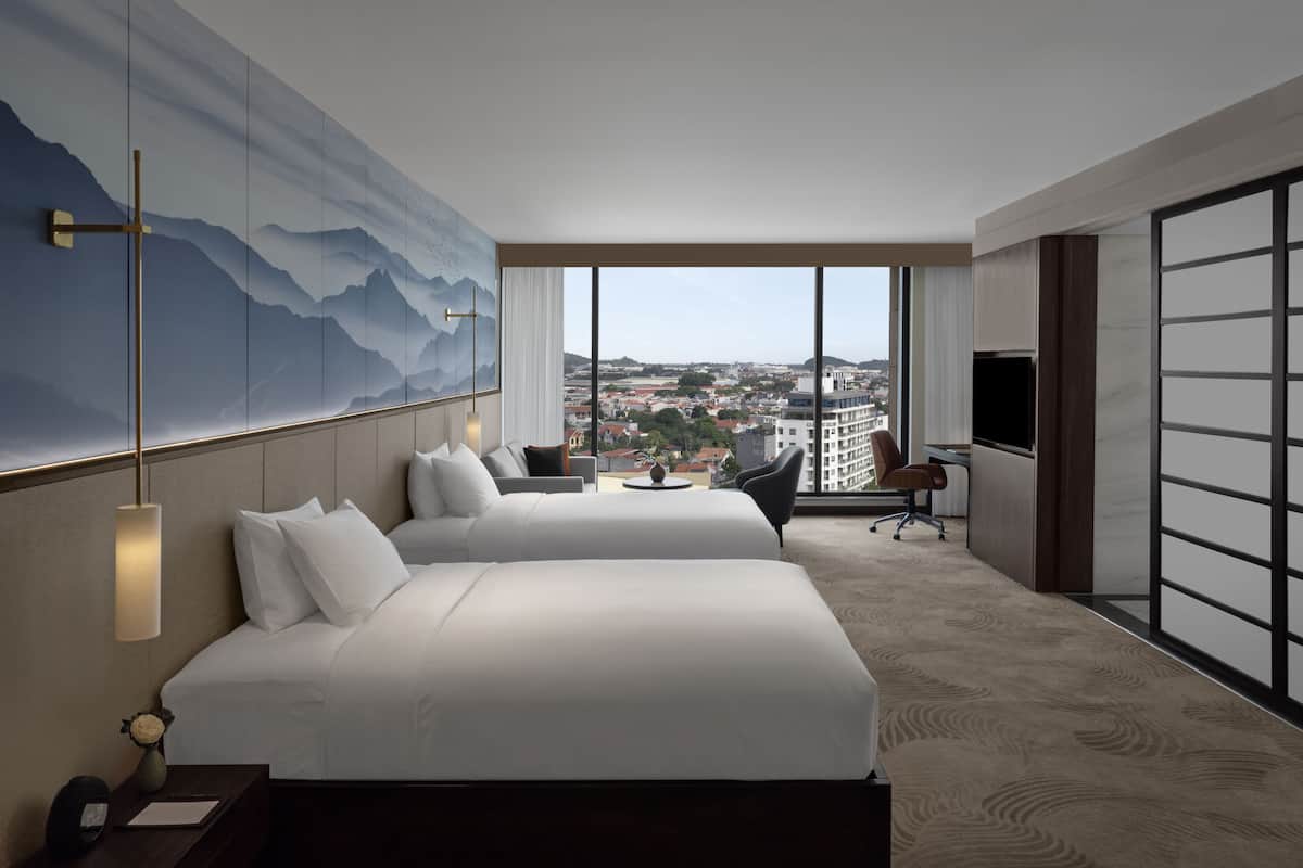 A hotel room with two beds, a desk, a chair, a TV, and a large window with a city view.