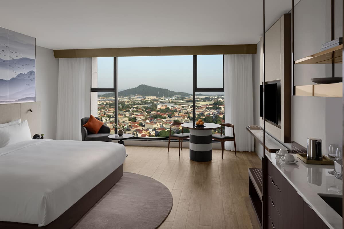 A hotel room with a large bed, a dining table, a TV, and a view of a cityscape.