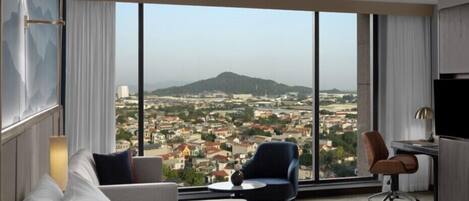 A hotel room with a large window offering a cityscape view, a bed, a desk, and a chair.