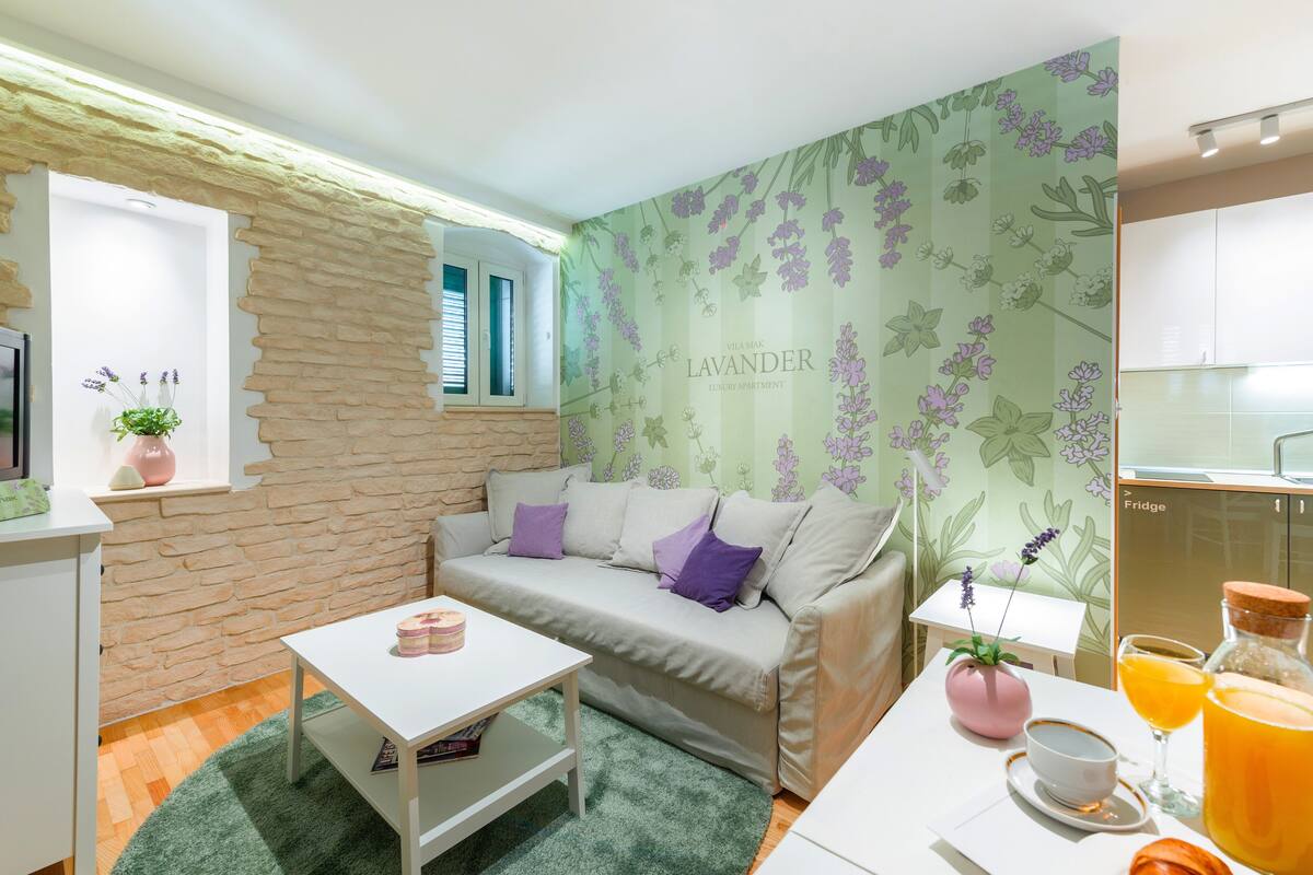 Ein gemütliches Wohnzimmer mit einer Couch, einem kleinen Tisch, einem Fenster mit Jalousien und einer Küchenzeile inklusive Kühlschrank.